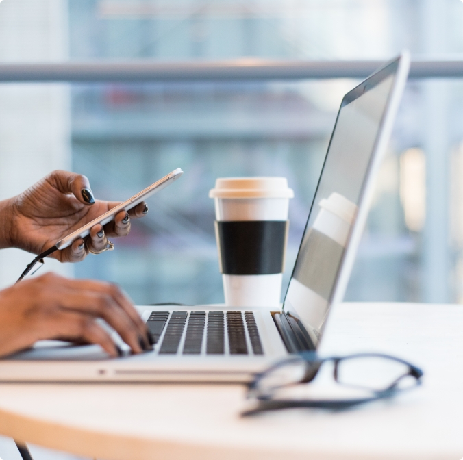 Woman working on laptop and using cell phone with coffee cup and glasses on desk