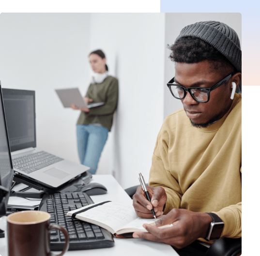 African student at desk writing in notebook with female student on laptop in background