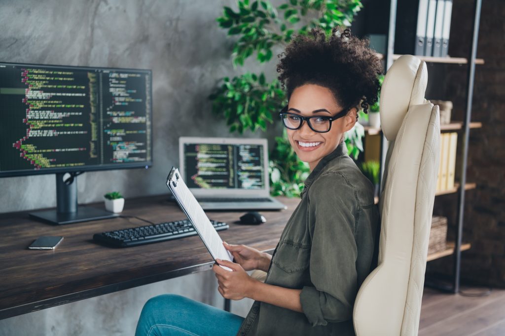 Female programmer witting a desk with dual screens and notebook